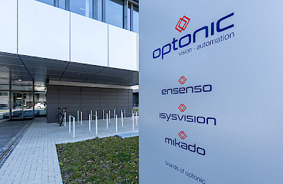 Silberne mit Optonic beschriftete Säule, vor dem grauen Optonic Gebäude mit den Marken Ensenso, Isysvision, mikado.