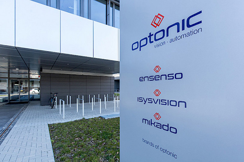 Silver column labeled Optonic, in front of the grey Optonic building with the brands Ensenso, Isysvision, mikado.