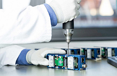 Person in a white coat and gloves assembling an assembly of the 3D Ensenso camera with a screwdriver.