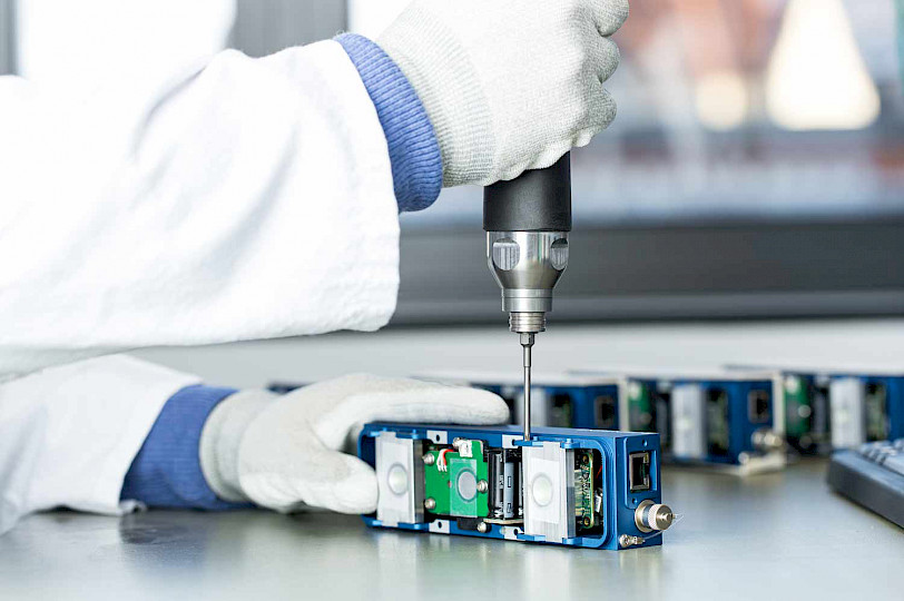 Person in a white coat and gloves assembling an assembly of the 3D Ensenso camera with a screwdriver.